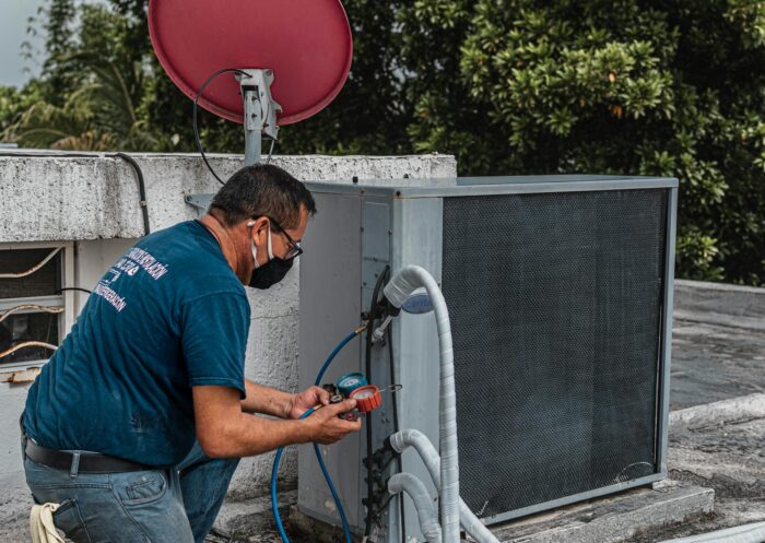 Technician repairing an air conditioner unit outdoors, wearing a facemask and using a manifold gauge.